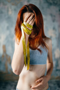  A skinny woman with her face covered by her hands, which are tied in measuring tape, representing weight loss and burning calories concept.