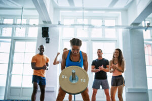 A group of young people cheering on their female friend lifting weights during a workout at the gym.