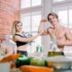 Fit couple in kitchen drinking smoothies, showcasing healthy eating and fitness lifestyle. Woman admires as man flexes biceps, highlighting proper nutrition's role in fitness.